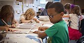 Skolmiljö där barn i sex-årsåldern sitter vid ett bord och ritar eller skriver. Barnet i förgrunden har en grön tröja och glasögon. Bilden är hämtad från en öppen bildbank.